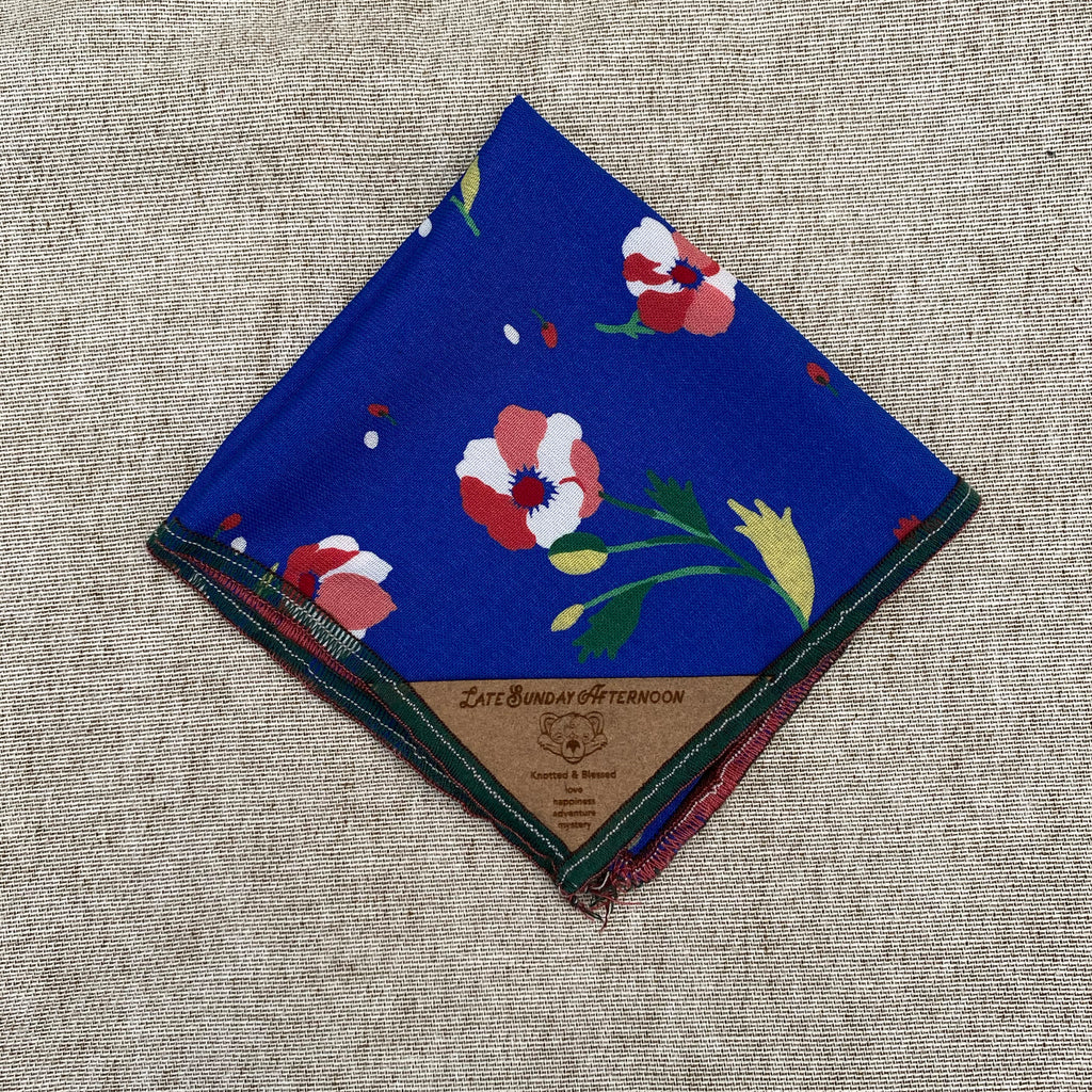 a folded pocket square with  red and white flowers with green and yellow stems on a blue background finished with a small brown tag on the corner