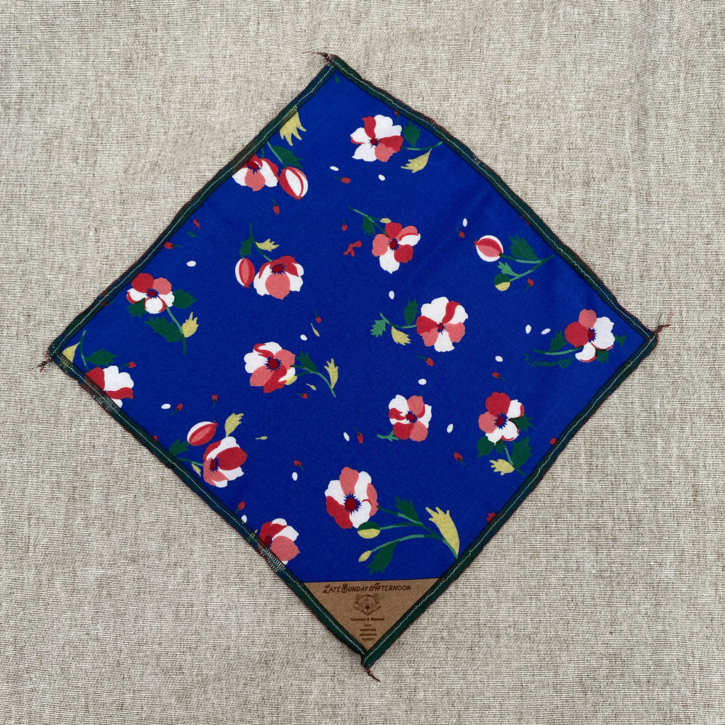 an open pocket square with red and white flowers with green and yellow stems on a blue background finished with a small brown tag on the corner